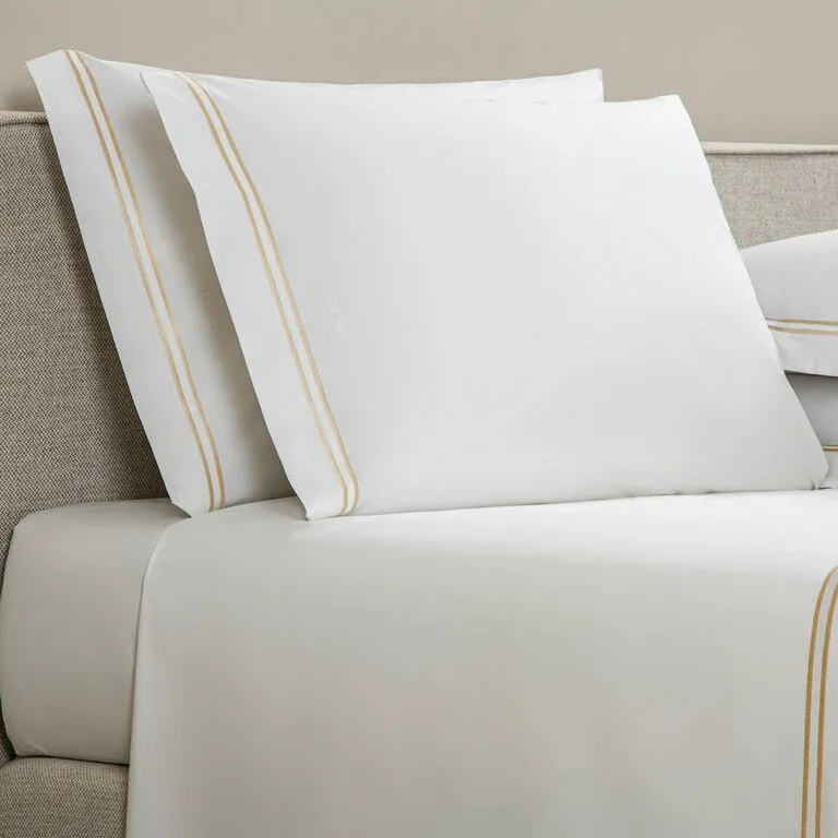The Frette Classic | Pillowcase Set by Frette, crafted from soft cotton percale with light brown embroidered stripes, adds a touch of luxury to your bed, perfectly complementing a beige upholstered frame.