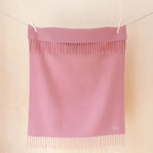 TBCo Lambswool Baby Blanket in Blush hanging from clothesline, displaying fringed edges against light wooden background