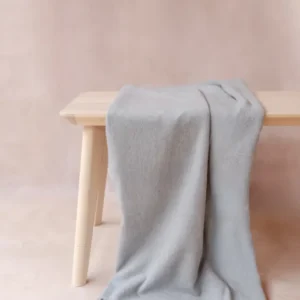 Cashmere Small Blanket in Light Grey Melange with fringe draped over bench, soft woven fabric by TBCo luxury collection