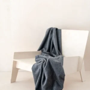 TBCo Lambswool Small Blanket in Charcoal Melange draped elegantly over white modern chair, showcasing soft texture and fringe detail