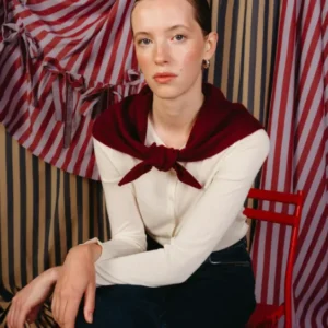Young woman models the TBCo Merino Wool Triangle Scarf in Burgundy, styled over her shoulders, complementing her cream top and dark jeans against a striped backdrop.