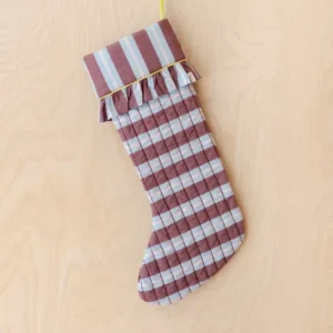 TBCo x Glassette Quilted Cotton Christmas Stocking in Berry Stripe features bold burgundy and blue stripes, ruffled cuff, and lime piping, hanging on a wood-grain wall.