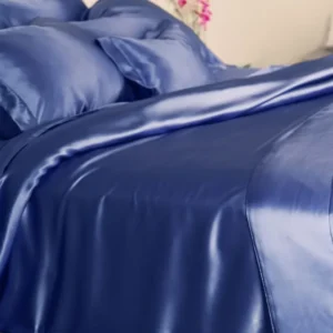 A softly lit bedroom features a partially rumpled Kumi Kookoon Classic Silk Twin Duvet Cover in shiny blue charmeuse silk, with matching pillowcases on the bed.