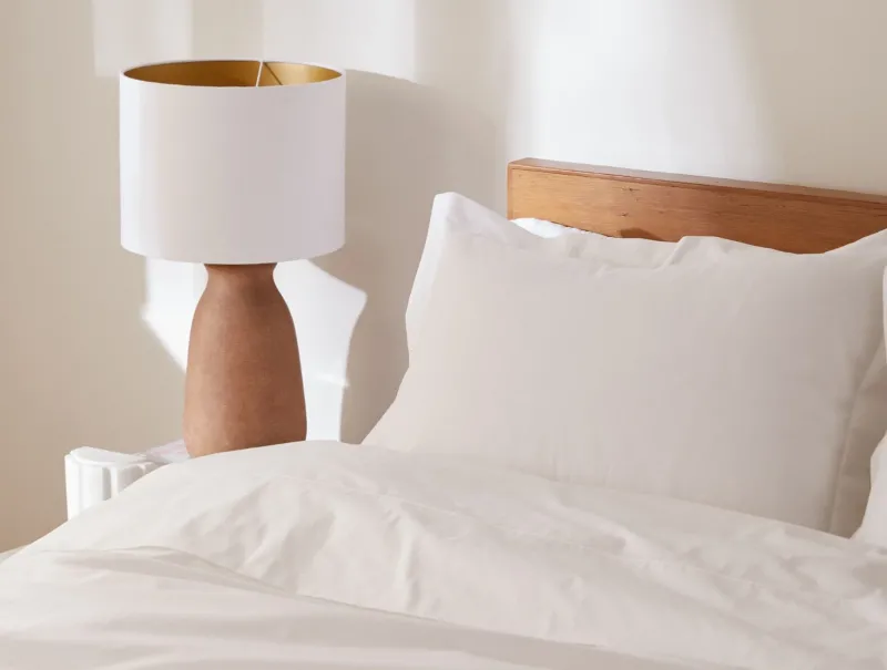 Coyuchi Heritage Percale pillowcases in white styled on bed beside warm wood lamp.